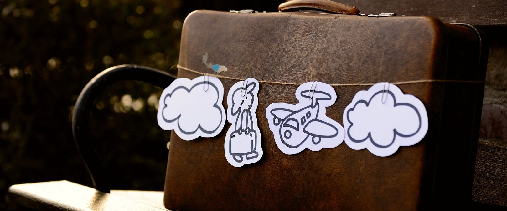 Ein brauner, alter Vintage-Koffer auf einer braunen Holzbank. Der Koffer ist mit einem Paketband umwickelt, auf dem verschiedene aus weißem Papier ausgeschnittene Reisesymbole hängen (Wolke, Flugzeug, Reisender).