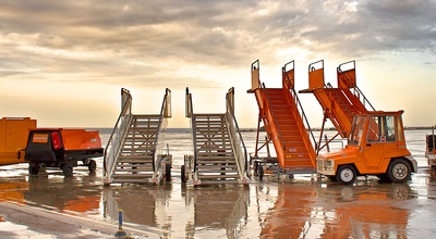 Ein Flughafen-Symbolbild: Es bildet einige Treppen und Rollfeld-Fahrzeuge ab, die Hauptfarbe ist Orange. Im Hintergrund Rollfeld und bewölkter Himmel in grau-orange Tönen.