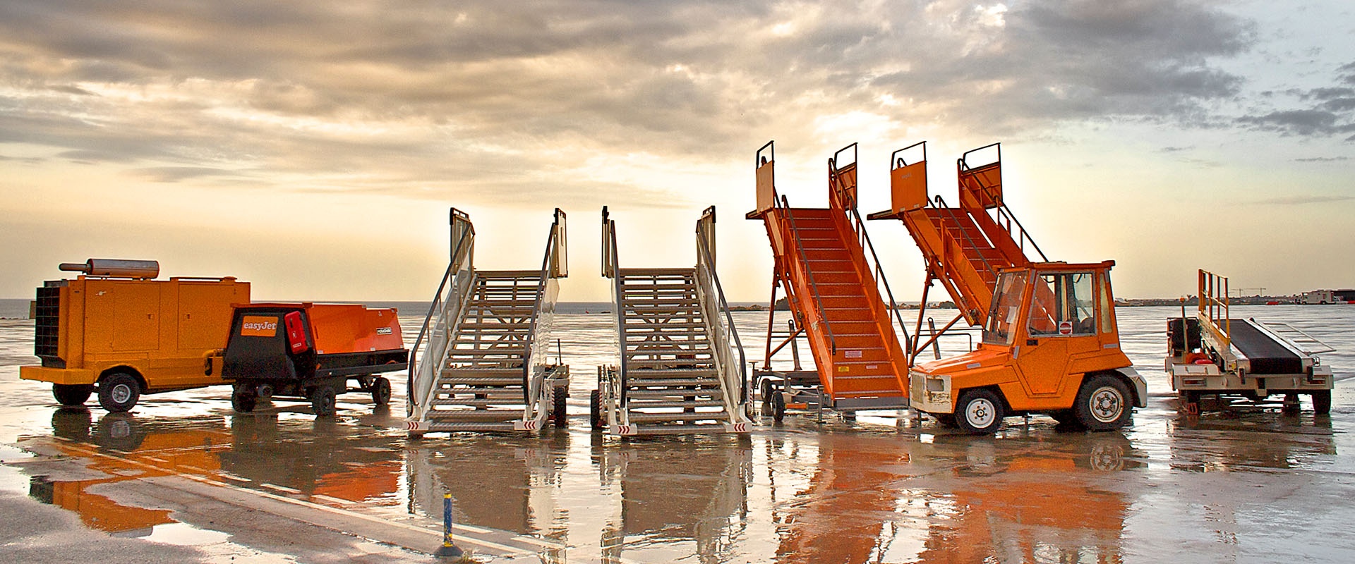 Ein Flughafen-Symbolbild: Es bildet einige Treppen und Rollfeld-Fahrzeuge ab, die Hauptfarbe ist Orange. Im Hintergrund Rollfeld und bewölkter Himmel in grau-orange Tönen.
