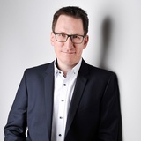 Ein lächelnder Mann im Business Look mit offenem Jackett, weißem Hemd mit schwarzgrau melierten Knöpfen und Brille, er steht vor einer weißen Wand mit leichtem Schattenwurf