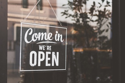 Ein Ausschnitt einer Glastür in der ein Schild mit der Aufschrift „Come in, we're open“ drauf steht, welches an eine Tafel erinnert, in der Glastür spiegeln sich einige Pflanzen und das gegenüberliegende Wohnhaus