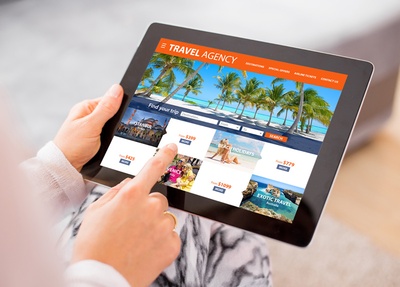 Die Nahaufnahme eines Tablets mit einer Reiseseite, das Tablet wird von einer Hand festgehalten, der Zeigefinger der anderen Hand zeigt auf ein Angebot