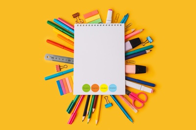 Ein weißer Block unter dem auf allen Seiten diverse bunte Büromaterialen hervorschauen (z.B. Buntstifte, Post-It-Streifen, Lineal, Schere oder Textmarker), das Ganze auf einem knallig orangem Hintergrund
