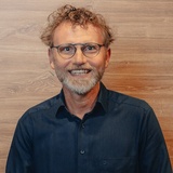 Ein lächelnder Mann mit freundlichem Blick, anthrazitfarbenem Hemd, Bart, Brille und einer dunkelblonden Kurzhaarfrisur vor einem hölzernen Hintergrund