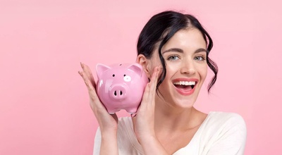 Eine Frau mit fröhlichem Gesichtsausdruck, dunklen Haaren und weißem T-Shirt hält ein rosa Sparschwein hoch. Der Hintergrund ist rosa.