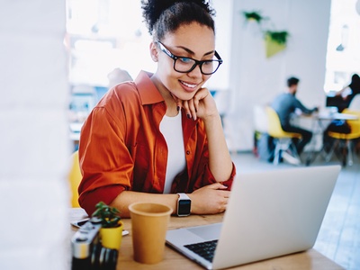 Eine lächelnde Frau sitzt an einem Tisch, vor ihr ein aufgeklapptes Notebook und ein Kaffeebecher, sie trägt ein weißes Top, darüber ein offenes oranges Hemd, eine SmartWatch und eine Hornbrille, ihre gelockten Haare sind zum Zopf zusammengebunden..