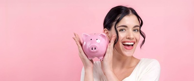 Eine Frau mit fröhlichem Gesichtsausdruck, dunklen Haaren und weißem T-Shirt hält ein rosa Sparschwein hoch. Der Hintergrund ist rosa.