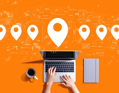 Aus Vogelperspektive sieht man ein offenes Notebook mit zwei Händen, eine Kaffeetasse und ein Notizbuch, alles auf knallig orangem Hintergrund, im oberen Bereich des Bilder sind in grafische Elemente, die an Ziel-Pins einer Navigation erinnern