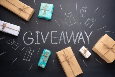 Auf schwarzem Tafelhintergrund steht mit weißer Kreide mittig „GIVEAWAY“, drum herum sind im Kreis Geschenke und ein paar Striche gemalt, außerdem liegen auf dem Bild verteilt mehrere kleine Päckchen in weiß, braun und Türkis mit Schnurschleifen