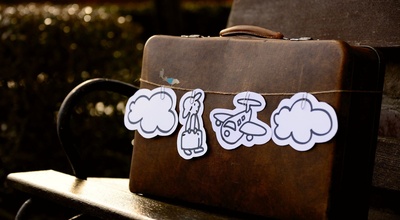 Ein brauner, alter Vintage-Koffer auf einer braunen Holzbank. Der Koffer ist mit einem Paketband umwickelt, auf dem verschiedene aus weißem Papier ausgeschnittene Reisesymbole hängen (Wolke, Flugzeug, Reisender).